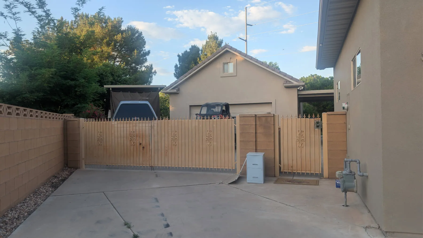 A faded tan wrought-iron double driveway gate with spear tops, closed across a concrete driveway between block pilasters